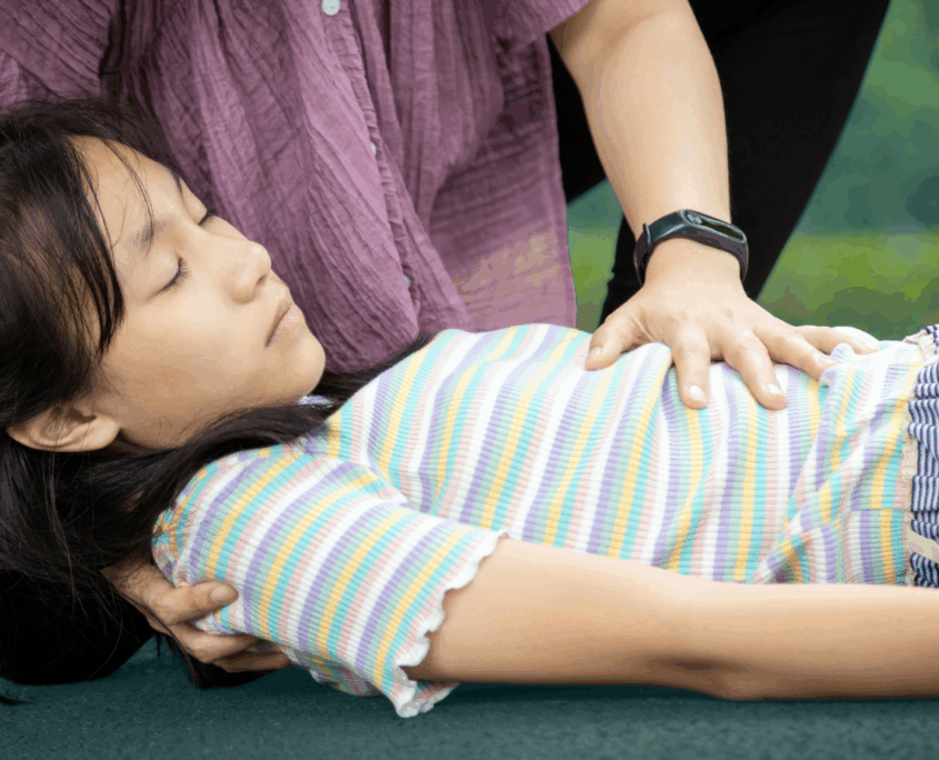 A girl about age 12 lies on the floor with her eyes closed. A woman puts her left hand on the girl's chest and cradles her head with her right hand.