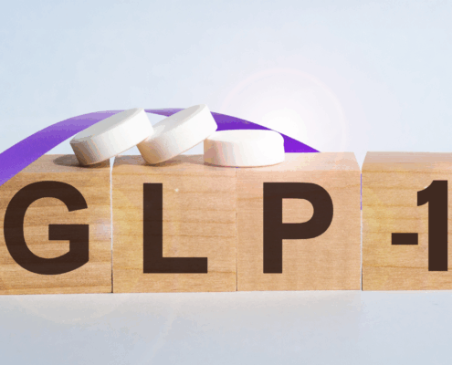 Four small brown blocks, similar to Scrabble tiles sit next to each other and spell out GLP-1. Three small white pills sit on top of the blocks and an image of a purple ribbons winds its way around the blocks.