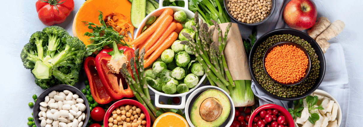 A display of brightly colored healthy foods includes a variety of vegetables and grains including carrots, Brussel sprouts, asparagus, half an avocado, red lentils and chickpeas.