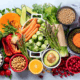 A display of brightly colored healthy foods includes a variety of vegetables and grains including carrots, Brussel sprouts, asparagus, half an avocado, red lentils and chickpeas.