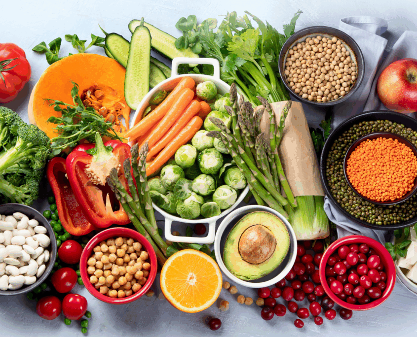 A display of brightly colored healthy foods includes a variety of vegetables and grains including carrots, Brussel sprouts, asparagus, half an avocado, red lentils and chickpeas.