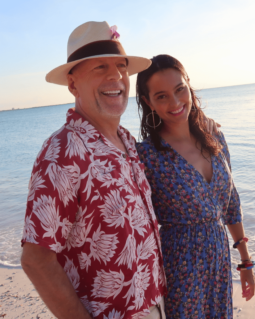 Bruce and Emma Willis stand next to the shore on a sandy beach while smiling at the camera.