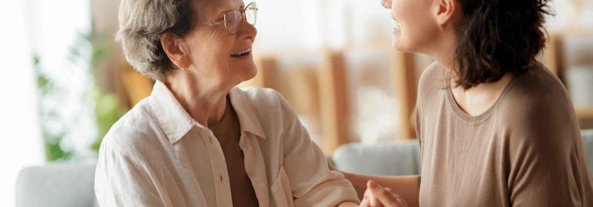A younger woman on the right faces an elderly woman while holding her hand. She appears to be trying to reassure the older woman.