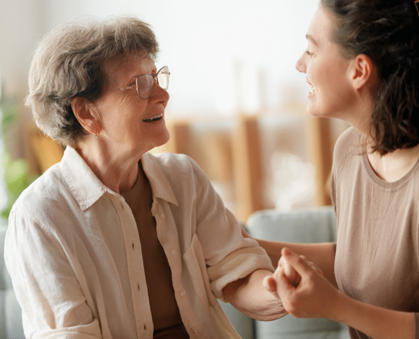 A younger woman on the right faces an elderly woman while holding her hand. She appears to be trying to reassure the older woman.