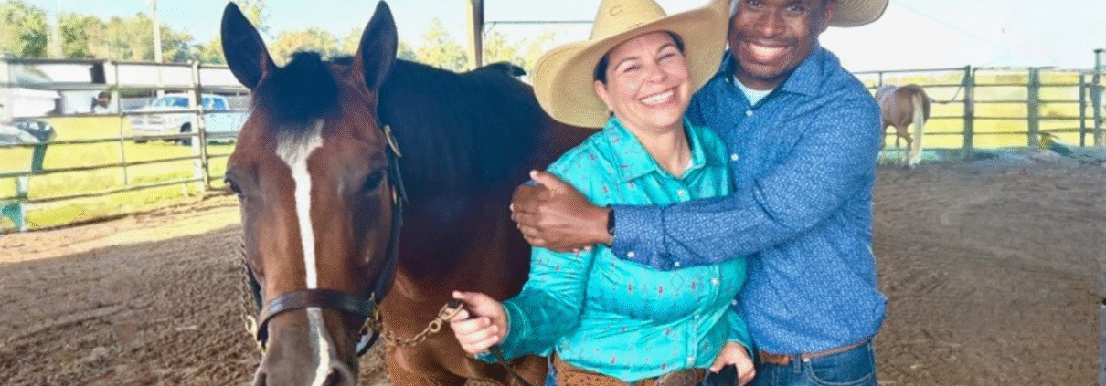Ernie Davis hugs his wife, Nikki, while wearing cowboy hats, jeans and button-down shirts and holding onto the reins of their brown horse standing next to them.