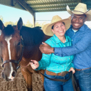 Ernie Davis hugs his wife, Nikki, while wearing cowboy hats, jeans and button-down shirts and holding onto the reins of their brown horse standing next to them.