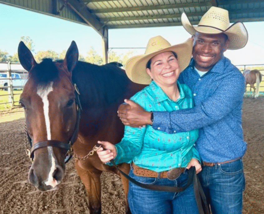 Ernie Davis hugs his wife, Nikki, while wearing cowboy hats, jeans and button-down shirts and holding onto the reins of their brown horse standing next to them.