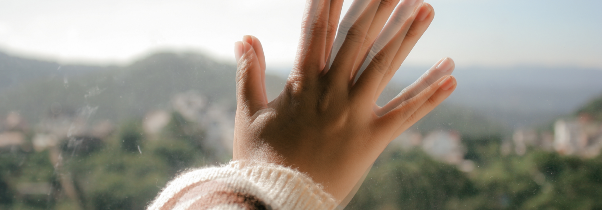 A white person's hand is pressed up against a glass window. The image of the hand is fuzzy and shows double the number of fingers. The outside view of nature and houses in the background is also blurry and hard to make out.