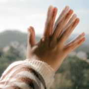 A white person's hand is pressed up against a glass window. The image of the hand is fuzzy and shows double the number of fingers. The outside view of nature and houses in the background is also blurry and hard to make out.