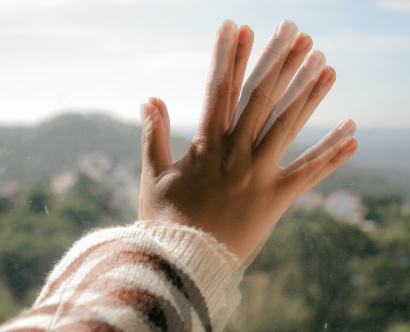 A white person's hand is pressed up against a glass window. The image of the hand is fuzzy and shows double the number of fingers. The outside view of nature and houses in the background is also blurry and hard to make out.