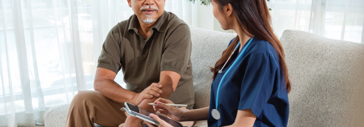 An older Asian man with gray hair and beard sits on a tan sofa talking to a young woman in blue healthcare scrubs. She holds a digital tablet and stylus while they talk.