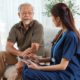 An older Asian man with gray hair and beard sits on a tan sofa talking to a young woman in blue healthcare scrubs. She holds a digital tablet and stylus while they talk.