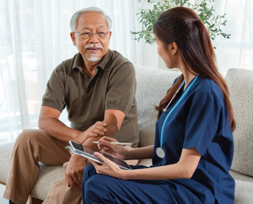 An older Asian man with gray hair and beard sits on a tan sofa talking to a young woman in blue healthcare scrubs. She holds a digital tablet and stylus while they talk.