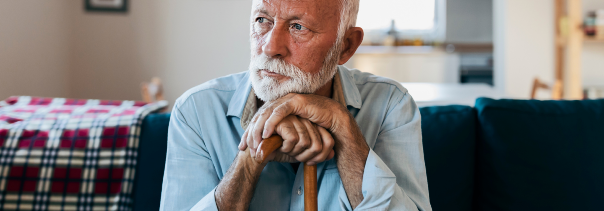 An older white man with white hair and groomed white beard and mustache sits on a couch. He rests both hands on the top of his cane in front of him and rests his head on his hands while he looks off into the distance.