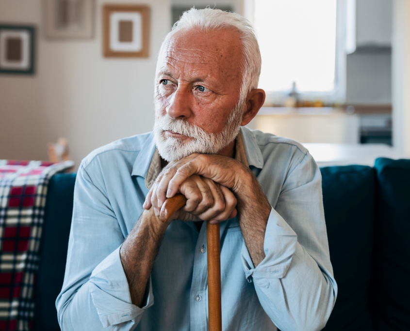 An older white man with white hair and groomed white beard and mustache sits on a couch. He rests both hands on the top of his cane in front of him and rests his head on his hands while he looks off into the distance.