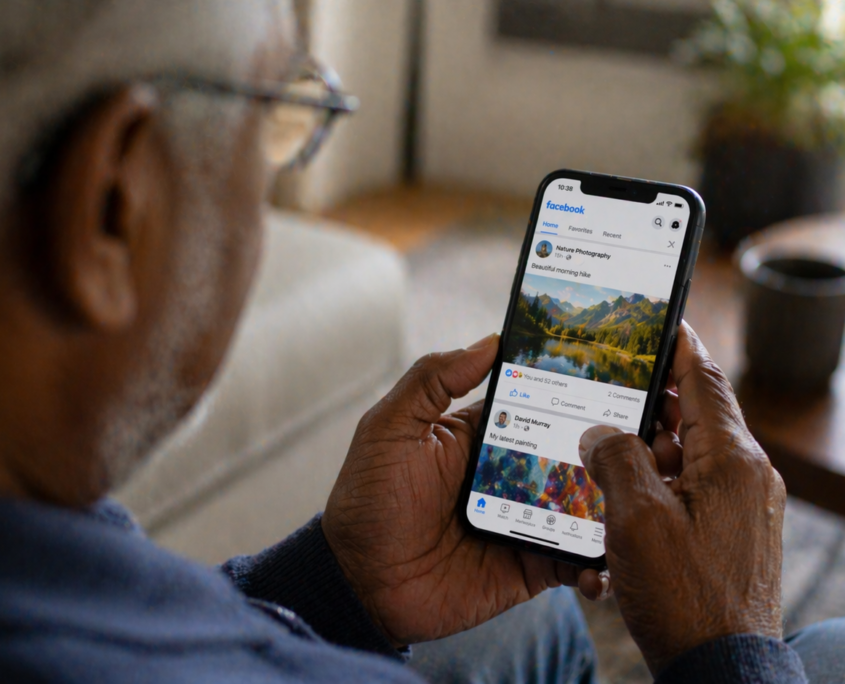 We see the blurry side of an older man from behind him as he looks down at a Facebook page on his smartphone he is holding in both hands.
