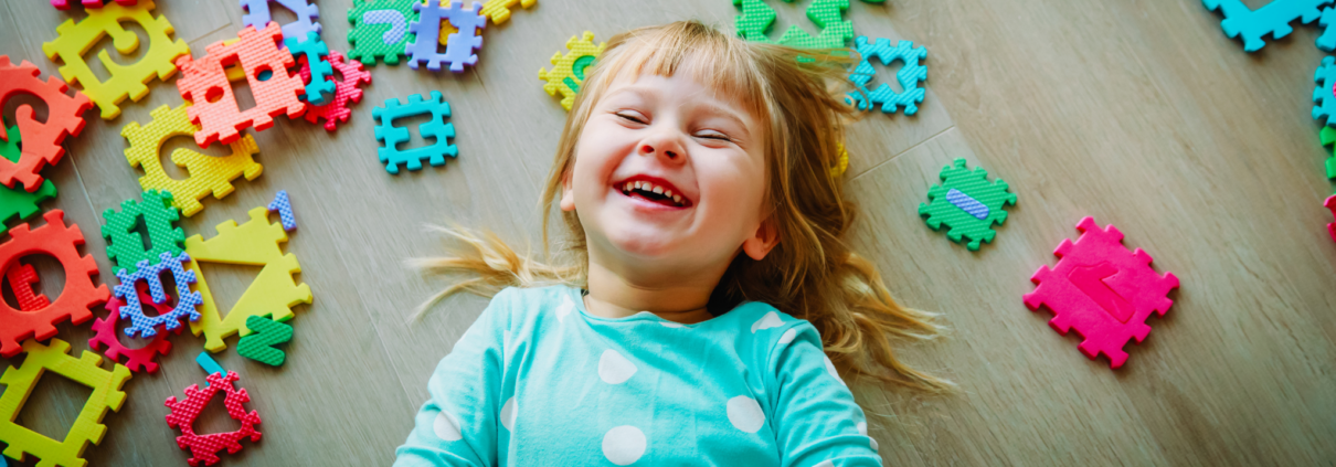 A preschool-age girl is laying on the floor and giggling. She has long blonde hair and is wearing a turquoise shirt with big white polka dots. She is surrounded by large puzzle pieces showing shapes, numbers and letters.