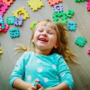 A preschool-age girl is laying on the floor and giggling. She has long blonde hair and is wearing a turquoise shirt with big white polka dots. She is surrounded by large puzzle pieces showing shapes, numbers and letters.