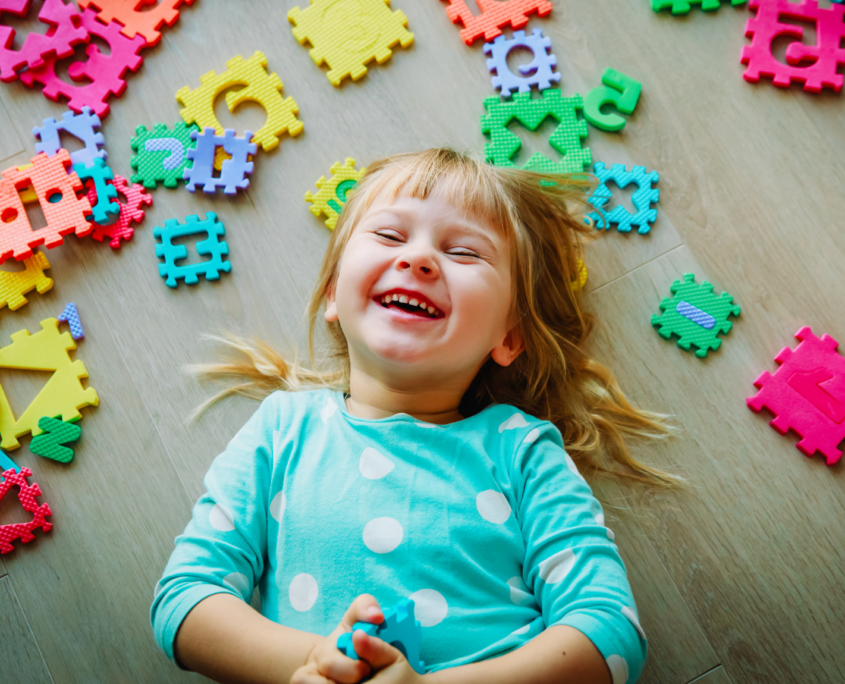 A preschool-age girl is laying on the floor and giggling. She has long blonde hair and is wearing a turquoise shirt with big white polka dots. She is surrounded by large puzzle pieces showing shapes, numbers and letters.