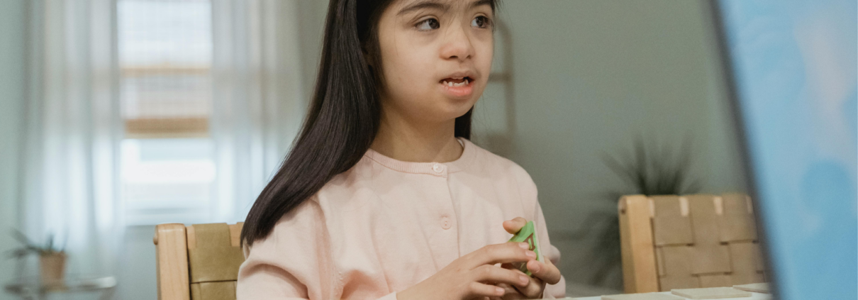 An autistic girl, about age 7, with long black hair is staring off into the distance while holding cards in her hands. A row of blurry unreadable cards are laid out on the table in front of her.