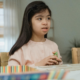 An autistic girl, about age 7, with long black hair is staring off into the distance while holding cards in her hands. A row of blurry unreadable cards are laid out on the table in front of her.
