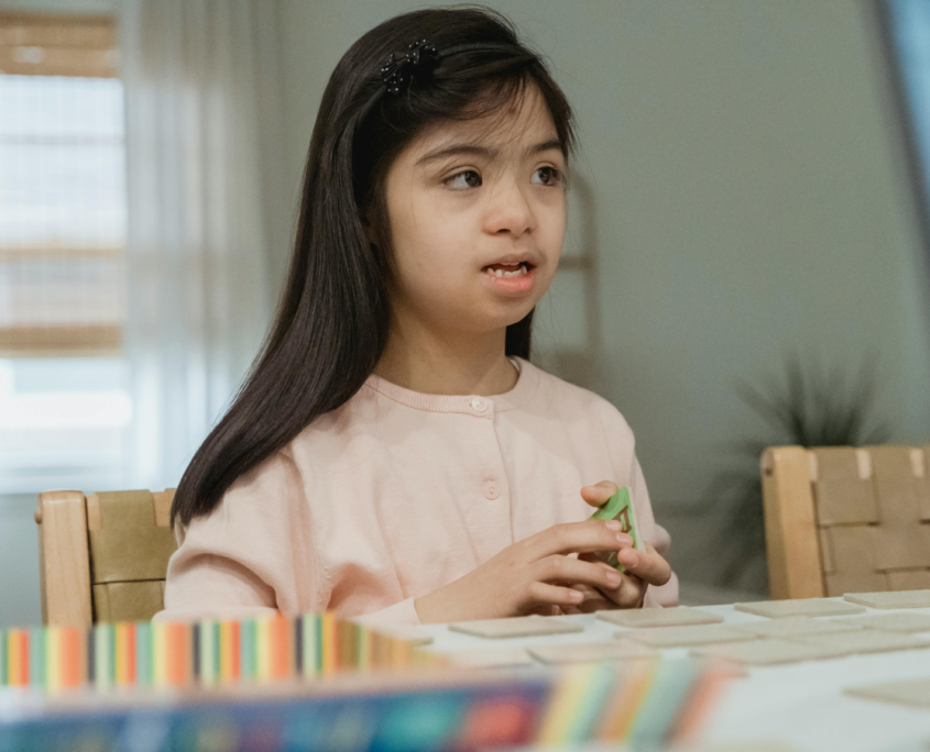 An autistic girl, about age 7, with long black hair is staring off into the distance while holding cards in her hands. A row of blurry unreadable cards are laid out on the table in front of her.