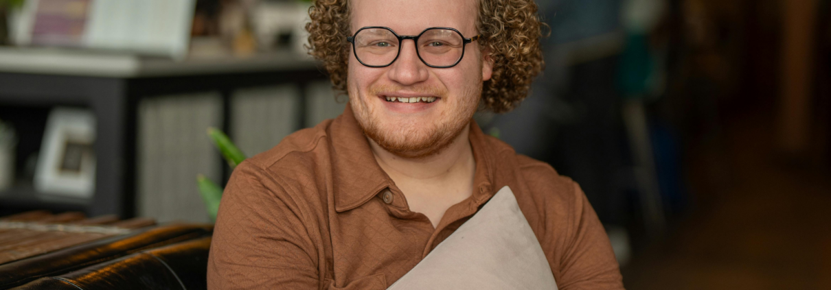 A young autistic man sits on a comfy chair and holds a pillow casually to his chest while smiling at the camera. He has tight curly reddish hair and a short red beard and is wearing glasses.