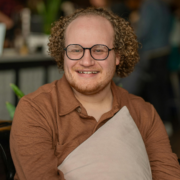 A young autistic man sits on a comfy chair and holds a pillow casually to his chest while smiling at the camera. He has tight curly reddish hair and a short red beard and is wearing glasses.