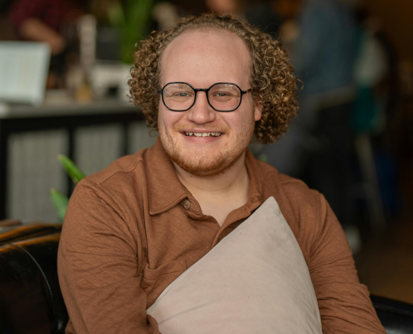 A young autistic man sits on a comfy chair and holds a pillow casually to his chest while smiling at the camera. He has tight curly reddish hair and a short red beard and is wearing glasses.