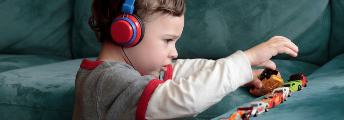 A preschool-age autistic boy is shown in profile playing with about 12 small cars lined up on a sofa cushion. The boy is wearing a white long-sleeve shirt with a gray T-shirt over it and wearing big blue and red headphones.