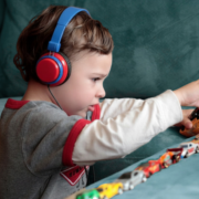 A preschool-age autistic boy is shown in profile playing with about 12 small cars lined up on a sofa cushion. The boy is wearing a white long-sleeve shirt with a gray T-shirt over it and wearing big blue and red headphones.