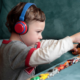 A preschool-age autistic boy is shown in profile playing with about 12 small cars lined up on a sofa cushion. The boy is wearing a white long-sleeve shirt with a gray T-shirt over it and wearing big blue and red headphones.