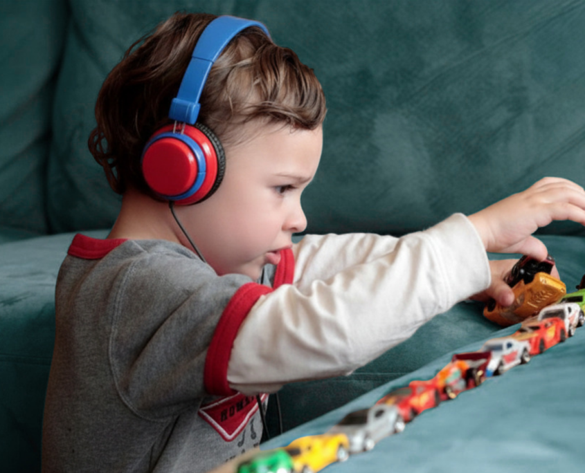 A preschool-age autistic boy is shown in profile playing with about 12 small cars lined up on a sofa cushion. The boy is wearing a white long-sleeve shirt with a gray T-shirt over it and wearing big blue and red headphones.