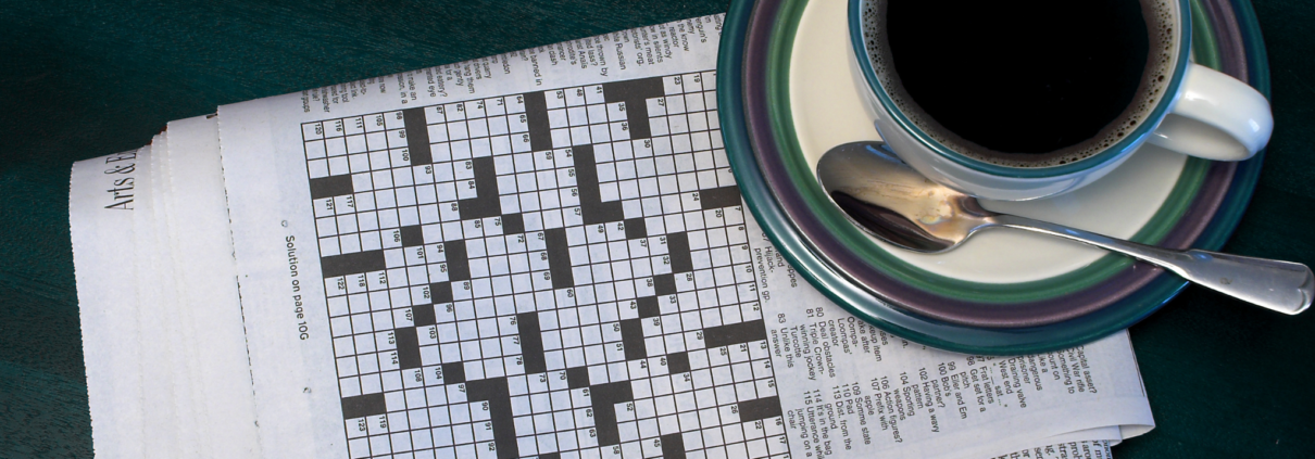 A newspaper is folded to the crossword puzzle, which has not been started yet. A tea cup filled with a dark liquid rests on a sauce, alongside a spoon.