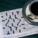 A newspaper is folded to the crossword puzzle, which has not been started yet. A tea cup filled with a dark liquid rests on a sauce, alongside a spoon.