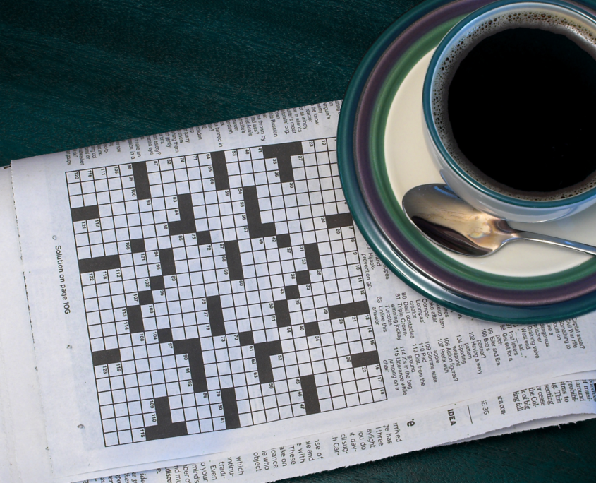 A newspaper is folded to the crossword puzzle, which has not been started yet. A tea cup filled with a dark liquid rests on a sauce, alongside a spoon.