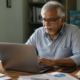 A man in his 60s with brown skin and gray hair, mustache and beard, wearing a light blue dress shirt and eyeglasses, sits at a desk in a home office, typing on a laptop.